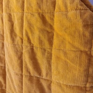 May include: Close-up of a mustard-yellow corduroy fabric with a quilted design. The fabric features vertical ridges and is divided into square sections by stitching. The colour is a warm, earthy tone.