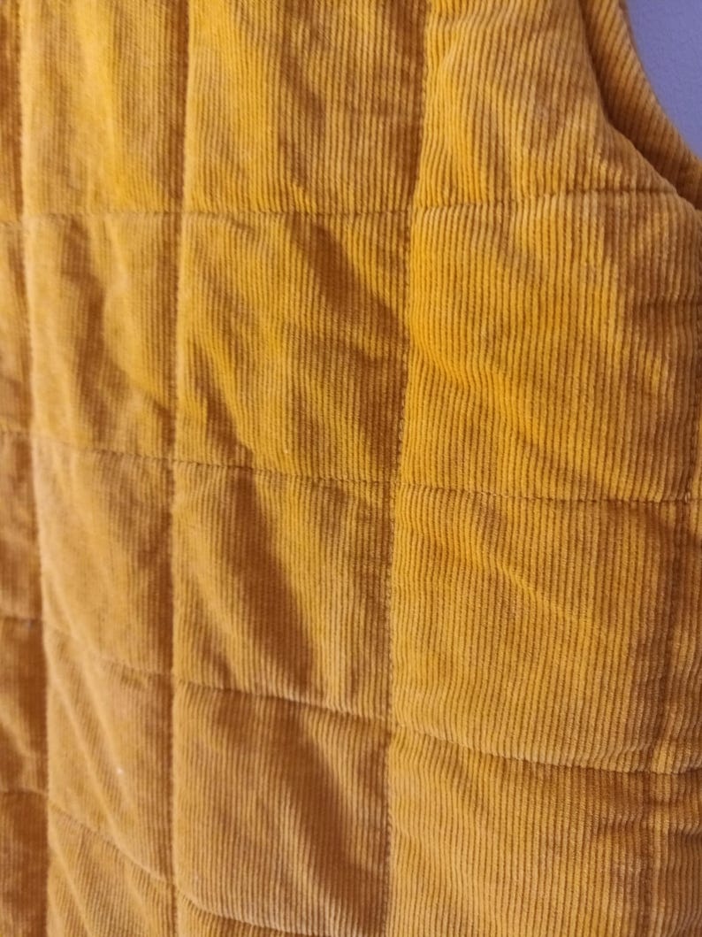 May include: Close-up of a mustard-yellow corduroy fabric with a quilted design. The fabric features vertical ridges and is divided into square sections by stitching. The colour is a warm, earthy tone.