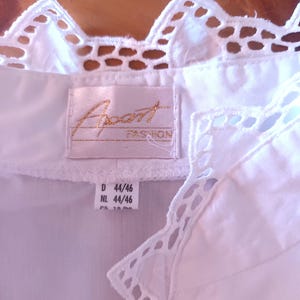 Pode incluir: Uma blusa branca com gola e acabamento em renda festonada decorativa. Uma etiqueta de tecido retangular est&aacute; costurada no decote com o texto Apart FASHION bordado em dourado. Uma etiqueta branca menor abaixo mostra os tamanhos D 44/46 e NL 44/46.
