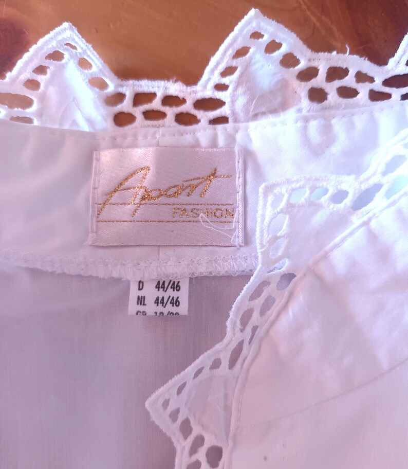 Pode incluir: Uma blusa branca com gola e acabamento em renda festonada decorativa. Uma etiqueta de tecido retangular est&aacute; costurada no decote com o texto Apart FASHION bordado em dourado. Uma etiqueta branca menor abaixo mostra os tamanhos D 44/46 e NL 44/46.