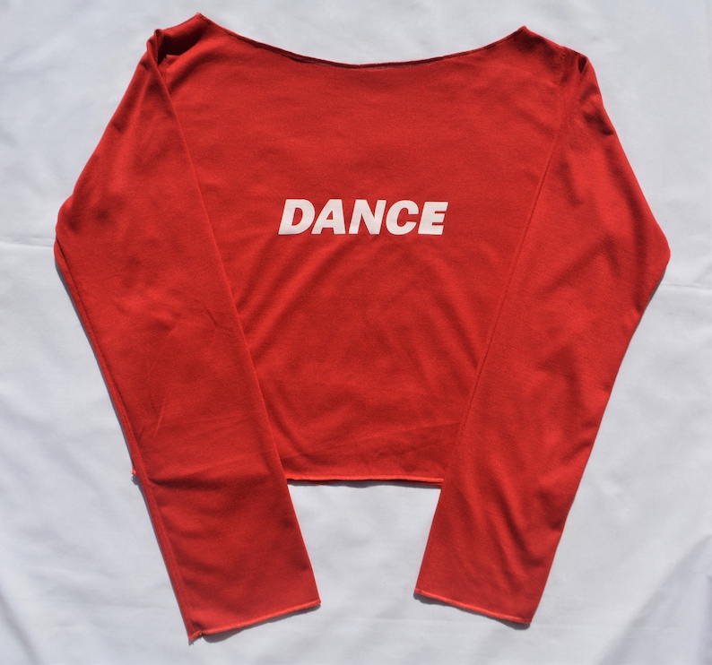 May include: Red long-sleeved shirt with a boat neck and the word "DANCE" printed in white on the front.