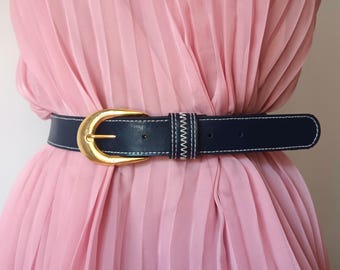 May include: A navy blue belt with a gold-colored buckle and white stitching. The belt is worn over a pink pleated dress. The buckle is a large, rounded shape. The belt is a classic accessory.