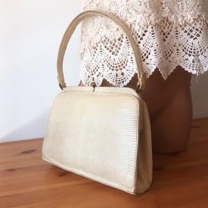 Könnte beinhalten: Cremefarbene Vintage-Handtasche mit einem strukturierten, reptilienartigen Muster. Die Tasche hat einen gebogenen Griff und einen Schnallenverschluss. Die Handtasche steht auf einer Holzoberfläche.