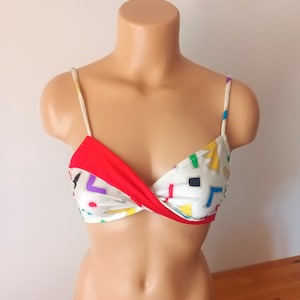 Peut inclure: Un haut de bikini blanc avec un motif géométrique rouge, jaune, bleu, vert et violet. Le haut a un détail de torsion à l'avant.