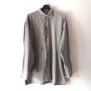Op de afbeelding: Grijs overhemd met lange mouwen, een knoopkraag en decoratieve borduursels op de borst. Het overhemd heeft een klassiek ontwerp met manchetten met knopen en een knoopsluiting. Het hangt aan een zwarte hanger tegen een witte achtergrond.