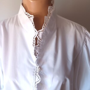 Pode incluir: Uma camisa branca de bot&otilde;es com gola alta e carcela frontal adornada com um intrincado acabamento em renda branca tipo ilh&oacute;. A pe&ccedil;a possui mangas curtas com detalhes em renda correspondentes nos punhos e um tecido liso e s&oacute;lido.