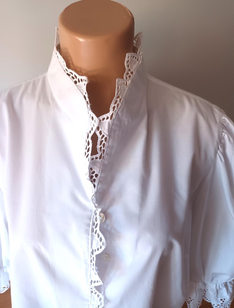 Pode incluir: Uma camisa branca de bot&otilde;es com gola alta e carcela frontal adornada com um intrincado acabamento em renda branca tipo ilh&oacute;. A pe&ccedil;a possui mangas curtas com detalhes em renda correspondentes nos punhos e um tecido liso e s&oacute;lido.