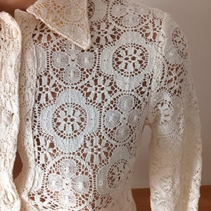 May include: A white crochet blouse with a long sleeve and a button-up front. The blouse has a delicate floral pattern.