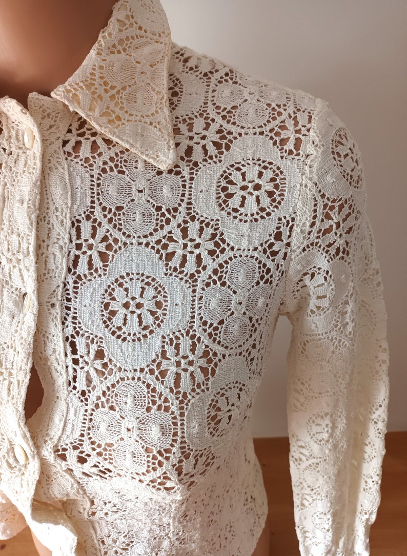 May include: A white crochet blouse with a long sleeve and a button-up front. The blouse has a delicate floral pattern.