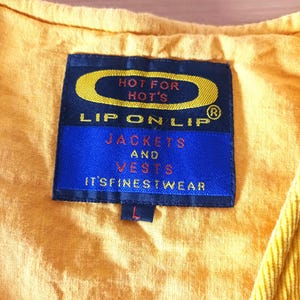 May include: A close-up of a yellow garment with a blue label. The label reads "HOT FOR HOT'S" and "LIP ON LIP" with the words "JACKETS AND VESTS" and "IT'S FINEST WEAR" below. The garment has a corduroy texture.