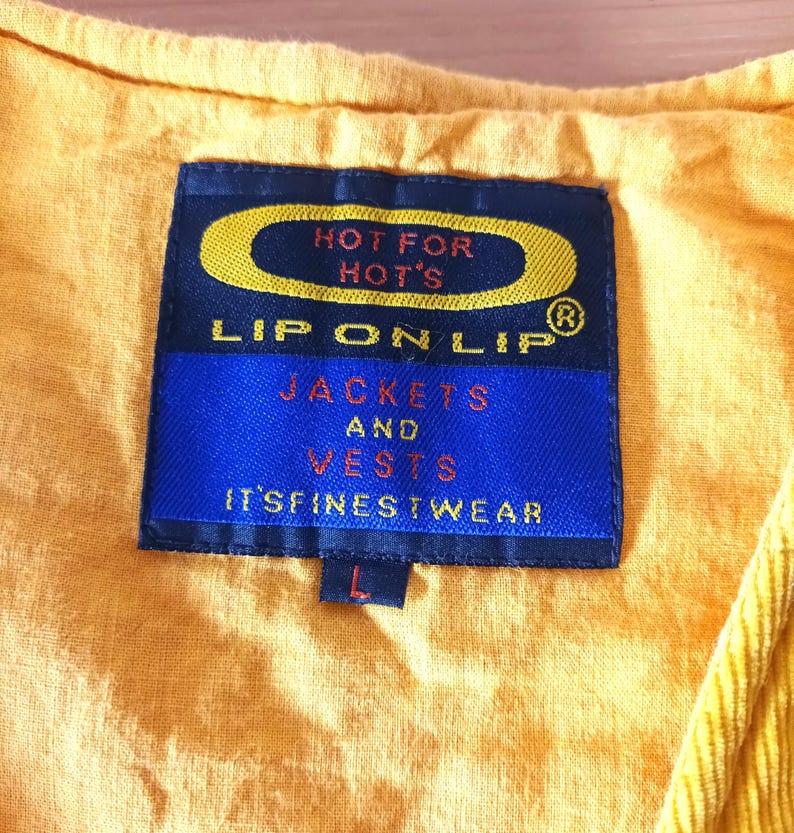 May include: A close-up of a yellow garment with a blue label. The label reads "HOT FOR HOT'S" and "LIP ON LIP" with the words "JACKETS AND VESTS" and "IT'S FINEST WEAR" below. The garment has a corduroy texture.