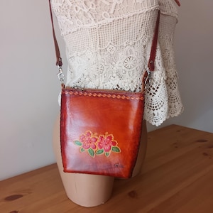 May include: A small brown leather crossbody bag with a floral design. The bag has a zipper closure and a long adjustable strap. The bag is made of genuine leather and is hand-stitched.