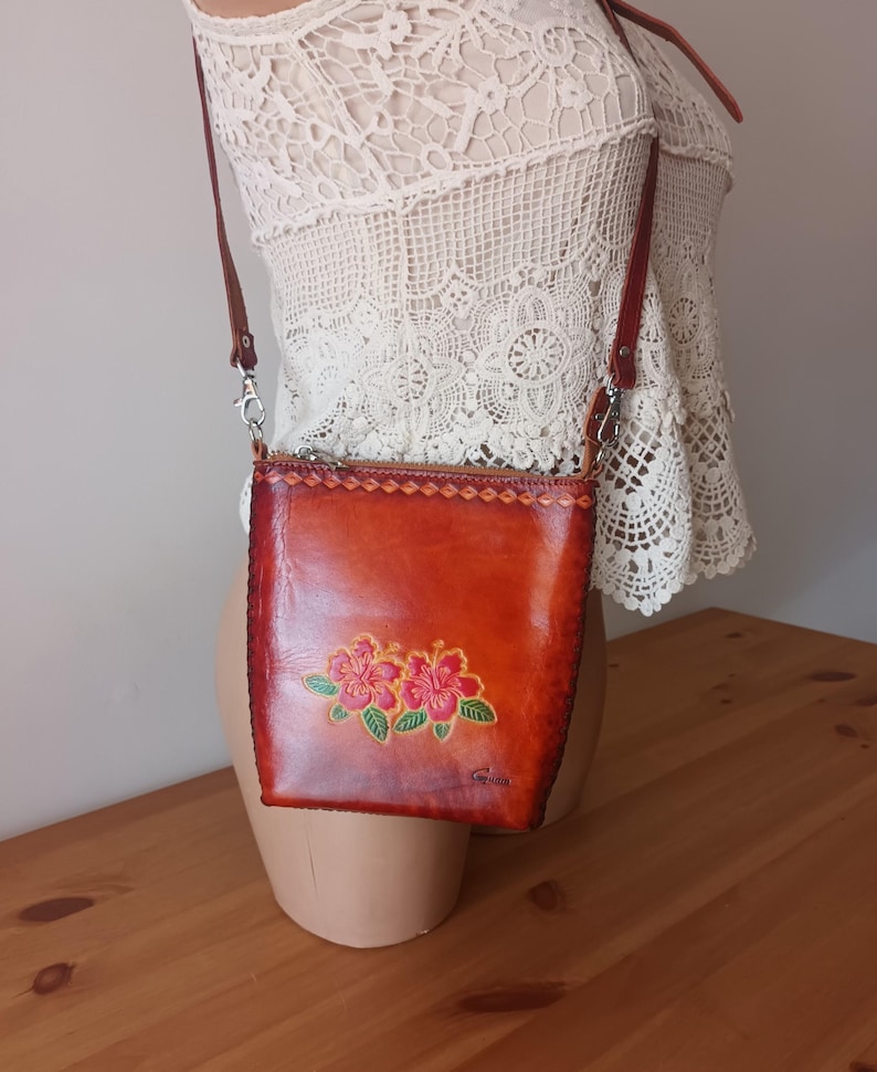 May include: A small brown leather crossbody bag with a floral design. The bag has a zipper closure and a long adjustable strap. The bag is made of genuine leather and is hand-stitched.