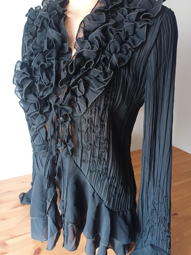Puede incluir: Blusa negra con cuello con volantes y mangas largas. La blusa presenta un escote en V y bordados decorativos en el corpi&ntilde;o y las mangas. La tela tiene una textura plisada y la parte inferior tiene un volante en capas.