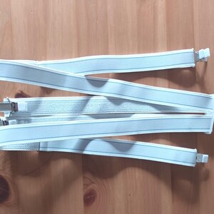 Puede incluir: Tirantes azul claro con clips metálicos plateados y correas elásticas blancas. Los tirantes están sobre una superficie de madera. Las correas tienen una sutil raya azul más clara en el centro.