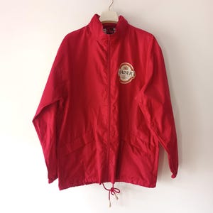 Puede incluir: Chaqueta roja con cremallera, cuello alto y dos bolsillos delanteros. La chaqueta tiene un parche con el logo "KAISER" en el pecho. La chaqueta está colgada sobre un fondo blanco.