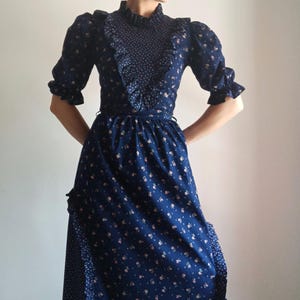Könnte beinhalten: Ein marineblaues Vintage-Kleid mit floralem Muster. Das Kleid hat einen hohen Rüschenkragen, kurze Puffärmel und eine taillierte Taille. Der lange Rock hat eine kontrastierende gemusterte Seite.