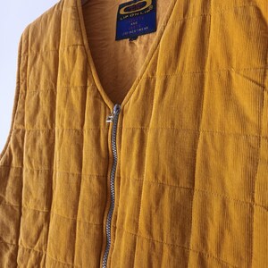 May include: Mustard yellow corduroy vest with a quilted design and a silver zip. The vest features a V-neck and a small label with the text "LIP ON LIP" and "JACKETS AND VESTS FINESTWEAR". It is hanging on a wooden hanger.