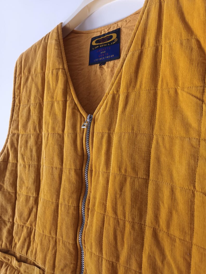 May include: Mustard yellow corduroy vest with a quilted design and a silver zip. The vest features a V-neck and a small label with the text "LIP ON LIP" and "JACKETS AND VESTS FINESTWEAR". It is hanging on a wooden hanger.
