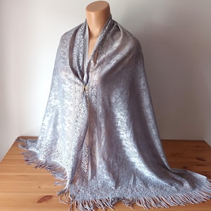 Può includere: Uno scialle grigio-argento con intricati motivi floreali e geometrici. Lo scialle è drappeggiato su un manichino, mostrando il suo design. I bordi sono rifiniti con frange. Una chiusura color oro fissa lo scialle al collo.