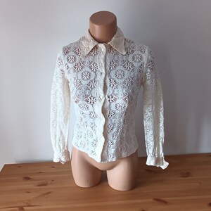 May include: A white lace blouse with long sleeves and a button-up front. The blouse has a delicate floral pattern and a collar.