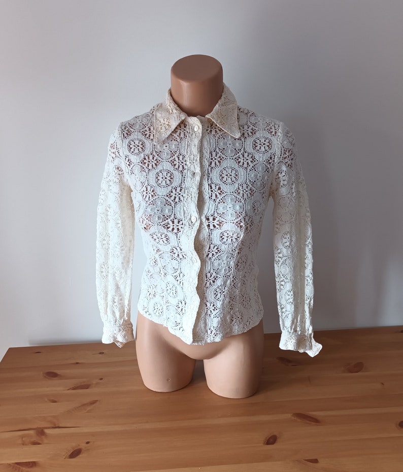 May include: A white lace blouse with long sleeves and a button-up front. The blouse has a delicate floral pattern and a collar.