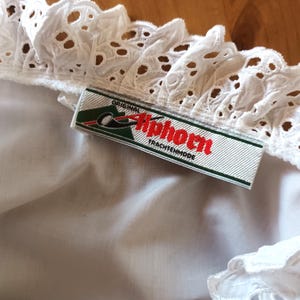 Puede incluir: Primer plano de una tela blanca con ojales y un borde decorativo. Una etiqueta con el texto "Original Tiphorn Trachtenmode" est&aacute; colocada sobre la tela. La etiqueta tiene un fondo verde y blanco con texto rojo.