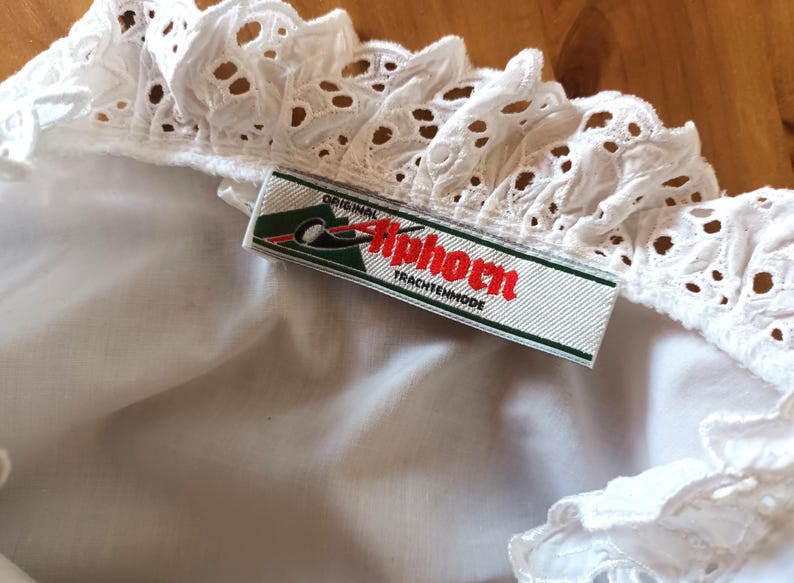 Puede incluir: Primer plano de una tela blanca con ojales y un borde decorativo. Una etiqueta con el texto "Original Tiphorn Trachtenmode" est&aacute; colocada sobre la tela. La etiqueta tiene un fondo verde y blanco con texto rojo.