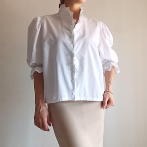 Pode incluir: Uma blusa branca com gola alta com acabamento em renda intrincada, mangas bufantes com punhos de renda franzidos e fecho frontal com bot&otilde;es. A blusa &eacute; combinada com uma saia lisa bege claro que chega abaixo da linha da cintura.