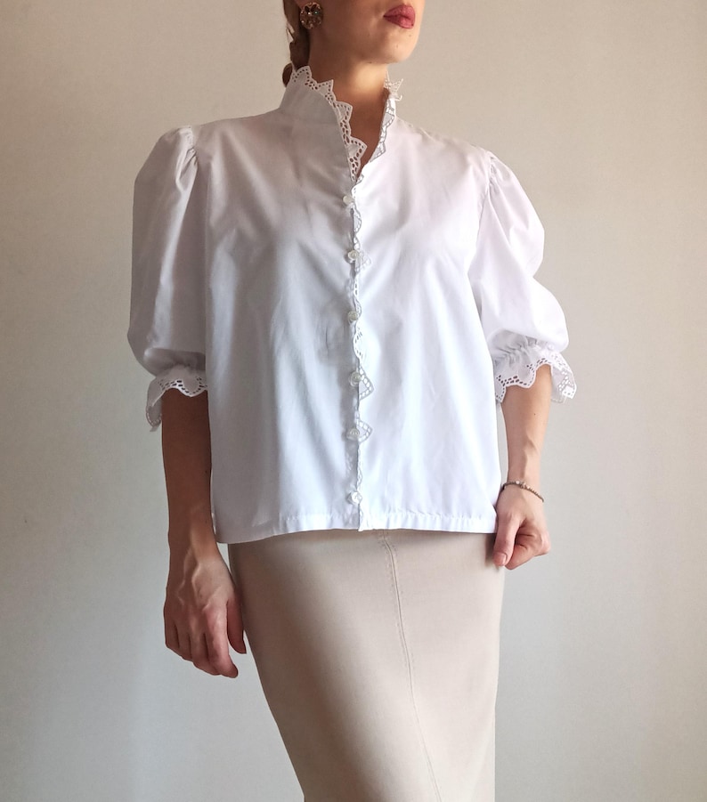 Pode incluir: Uma blusa branca com gola alta com acabamento em renda intrincada, mangas bufantes com punhos de renda franzidos e fecho frontal com bot&otilde;es. A blusa &eacute; combinada com uma saia lisa bege claro que chega abaixo da linha da cintura.