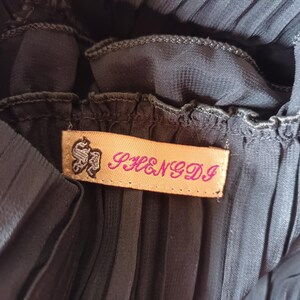 Puede incluir: Primer plano de una tela plisada gris oscuro. Una etiqueta rectangular con el texto "SHENGDI" en letras rosas est&aacute; cosida a la tela. La etiqueta tiene un emblema decorativo en negro.