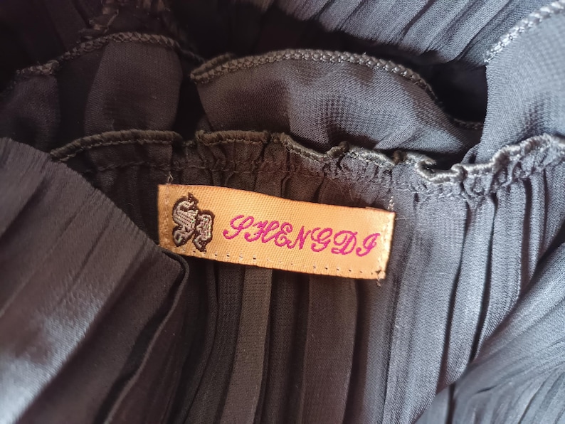 Puede incluir: Primer plano de una tela plisada gris oscuro. Una etiqueta rectangular con el texto "SHENGDI" en letras rosas est&aacute; cosida a la tela. La etiqueta tiene un emblema decorativo en negro.