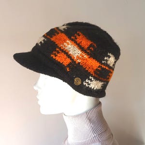 May include: A black knitted hat with a short peak, patterned with orange and white geometric shapes. A gold-coloured spiral button is on the side. The hat is displayed on a mannequin head.