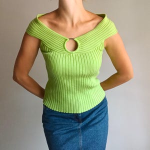 Pode incluir: Um top de malha canelada verde limão com um design único de ombro a ombro. O top apresenta um detalhe metálico circular no decote e uma silhueta ajustada. A modelo está usando uma saia jeans. Uma adição elegante a qualquer guarda-roupa.