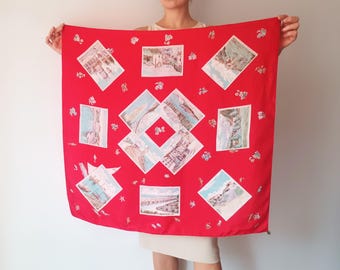 May include: A vibrant red square scarf featuring a repeating pattern of framed landscape illustrations. The illustrations are in shades of blue, white, and tan. Small floral accents are scattered across the scarf's surface. The scarf is held up by a person.