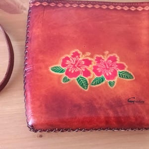May include: A small, brown leather purse with a floral design. The purse has a strap and a metal clasp. The design features two red hibiscus flowers with green leaves. The purse is labeled "Guam".