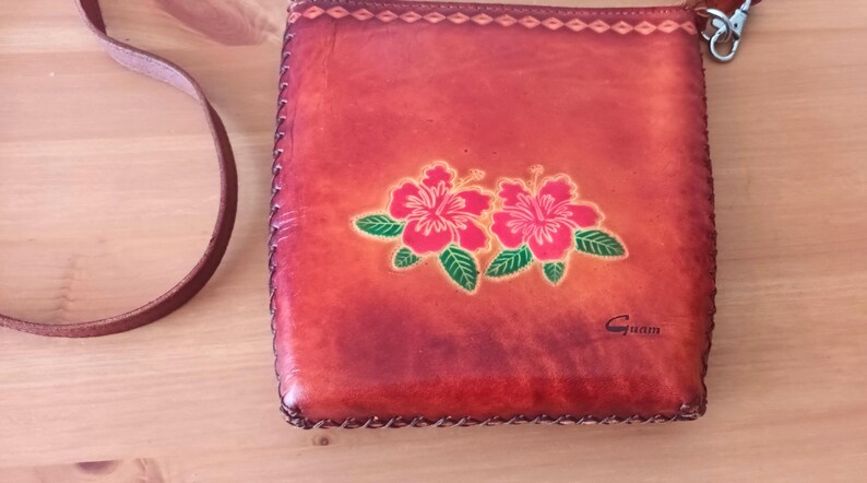 May include: A small, brown leather purse with a floral design. The purse has a strap and a metal clasp. The design features two red hibiscus flowers with green leaves. The purse is labeled "Guam".