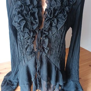 Puede incluir: Blusa negra con ribete de volantes y mangas largas. La blusa presenta un escote en V profundo y un cierre de lazo. La tela tiene una textura plisada y detalles bordados.