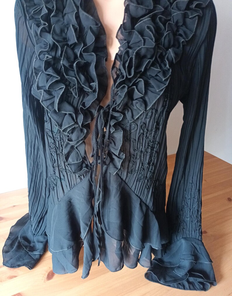 Puede incluir: Blusa negra con ribete de volantes y mangas largas. La blusa presenta un escote en V profundo y un cierre de lazo. La tela tiene una textura plisada y detalles bordados.