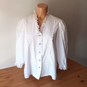Puede incluir: Blusa blanca con cuello en V y mangas largas. La blusa presenta intrincados detalles de encaje a lo largo del escote, en la parte delantera y en las mangas. Tiene una botonadura frontal y se exhibe en un maniqu&iacute;.