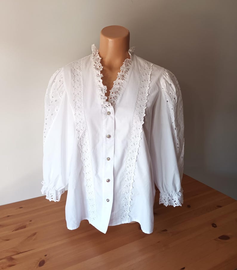 Puede incluir: Blusa blanca con cuello en V y mangas largas. La blusa presenta intrincados detalles de encaje a lo largo del escote, en la parte delantera y en las mangas. Tiene una botonadura frontal y se exhibe en un maniqu&iacute;.