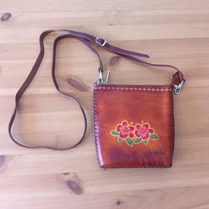 May include: A brown leather crossbody bag with a floral design. The bag has a zipper closure and a long adjustable strap. The bag is decorated with three red hibiscus flowers with green leaves.