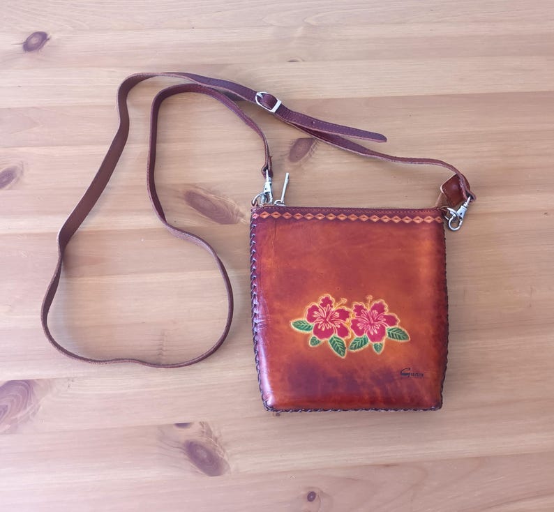May include: A brown leather crossbody bag with a floral design. The bag has a zipper closure and a long adjustable strap. The bag is decorated with three red hibiscus flowers with green leaves.