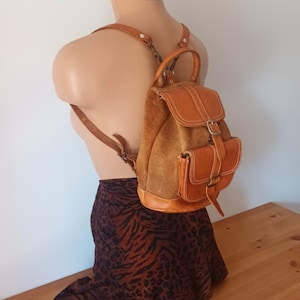 Puede incluir: Una mochila de cuero marrón con un panel frontal de gamuza y un cierre de hebilla. La mochila tiene un pequeño bolsillo en la parte delantera y correas ajustables.