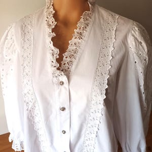 Puede incluir: Blusa blanca con cuello con volantes y ribete de encaje calado. La camisa de manga larga presenta botones decorativos en la parte delantera y detalles calados en las mangas. Prenda de inspiraci&oacute;n vintage.