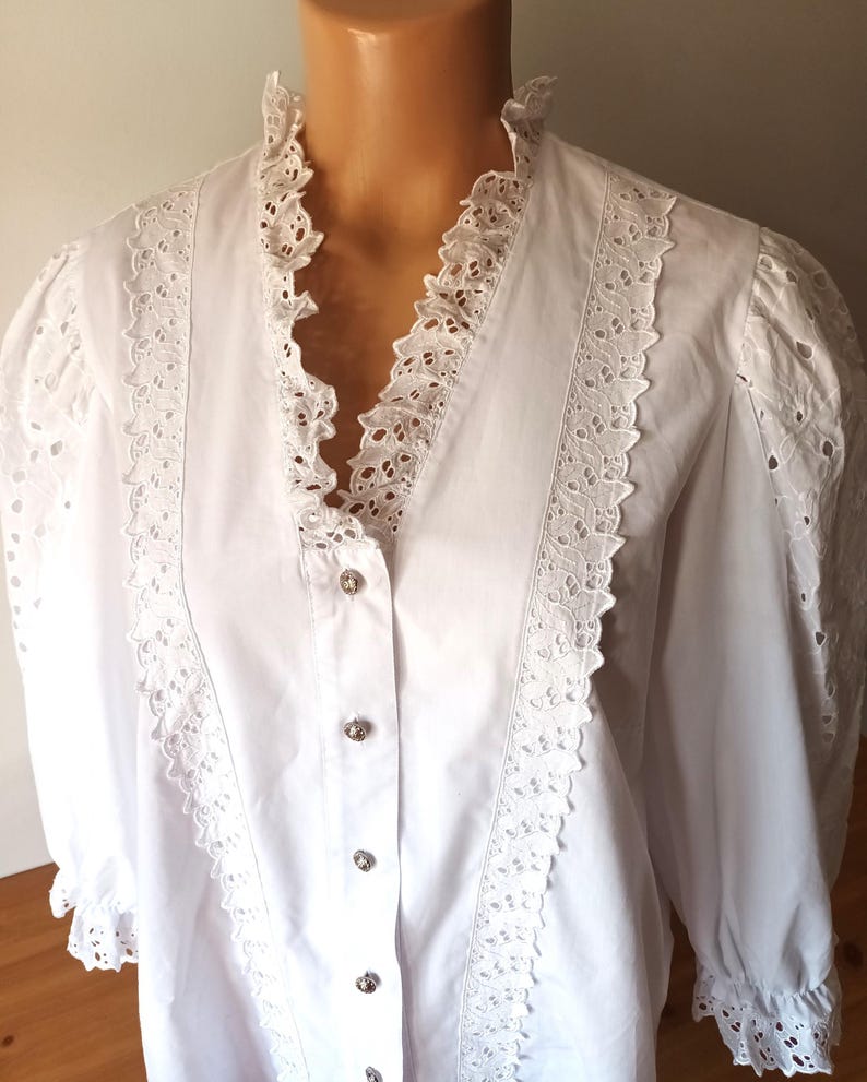 Puede incluir: Blusa blanca con cuello con volantes y ribete de encaje calado. La camisa de manga larga presenta botones decorativos en la parte delantera y detalles calados en las mangas. Prenda de inspiraci&oacute;n vintage.