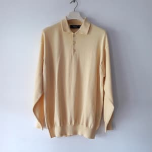 Puede incluir: Un polo de manga larga amarillo pálido con cuello y tres botones. La camisa está hecha de un material de punto suave y cuelga de una percha blanca sobre un fondo blanco. Los puños y el dobladillo son acanalados.