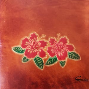 May include: A brown leather bag with a floral design featuring two red hibiscus flowers with yellow centers and green leaves. The bag has a stitched border and the word "Guam" is embossed on the bottom right corner.