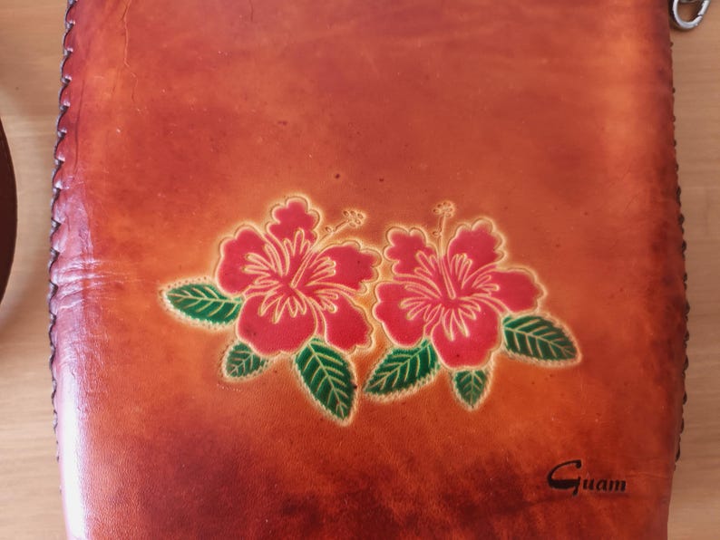 May include: A brown leather bag with a floral design featuring two red hibiscus flowers with yellow centers and green leaves. The bag has a stitched border and the word "Guam" is embossed on the bottom right corner.