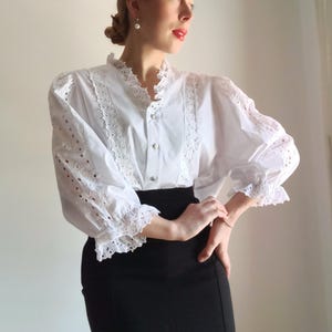 Puede incluir: Blusa blanca de ojal con cuello alto y mangas largas. La blusa presenta intrincados detalles de encaje a lo largo del cuello, la tapeta y las mangas. Las mangas tienen un pu&ntilde;o recogido y con volantes. La blusa se combina con una falda negra.